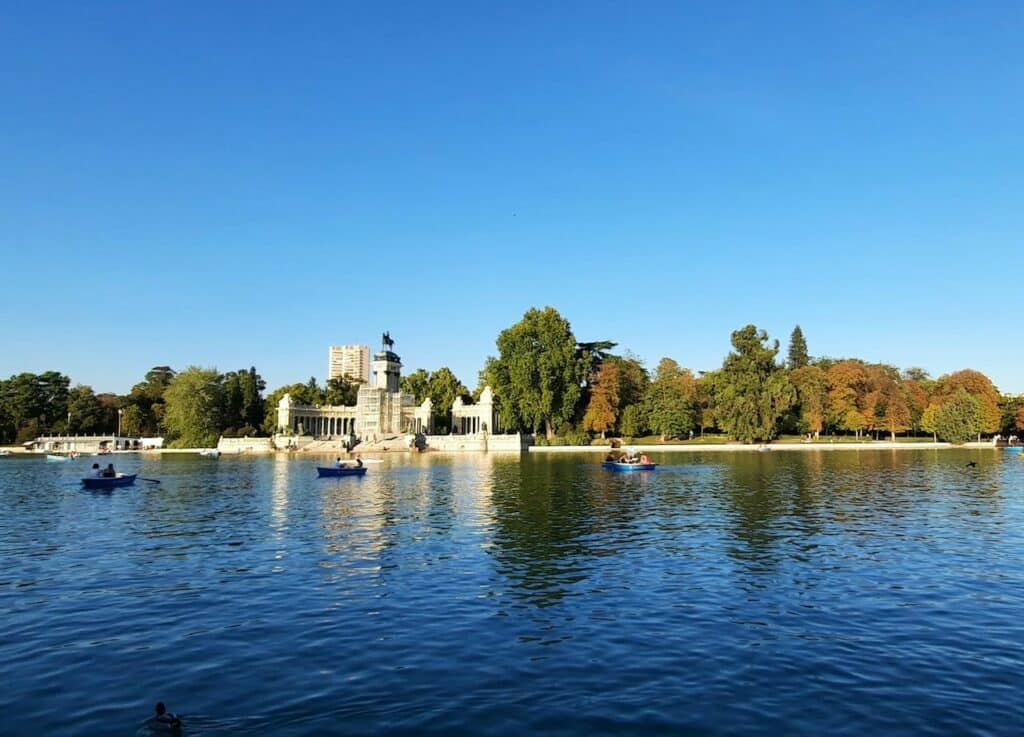 Vista do Parque do Retiro, com um lago e pessoas a andar de barco a remos, sendo que ao fundo há árvores e uma construção com estátua, o Monumento a Afonso XII