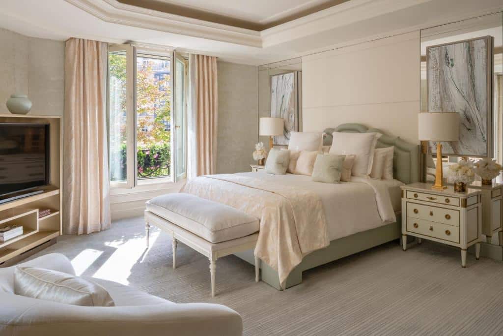 Um quarto bem iluminado do Four Seasons Hotel George V Paris com uma cama grande, banco, TV, poltrona, mesas de cabeceira e janelas grandes com cortinas claras.