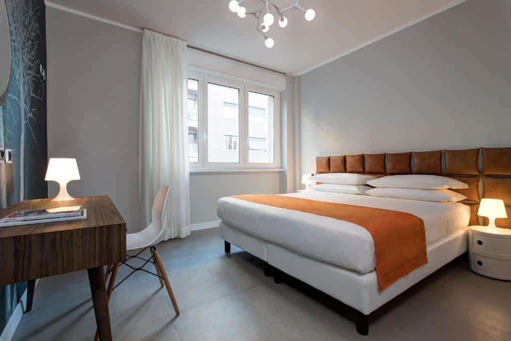 quarto do Noba Hotel e Residenze, um dos hotéis baratos em Roma, com cama de casal com pormenores laranja e branco, lustre de bolas, mesa de madeira e cadeira moderna, mesas de cabeceira e candeeiro de ambos os lados da cama, televisão em frente e janela com cortinados claros