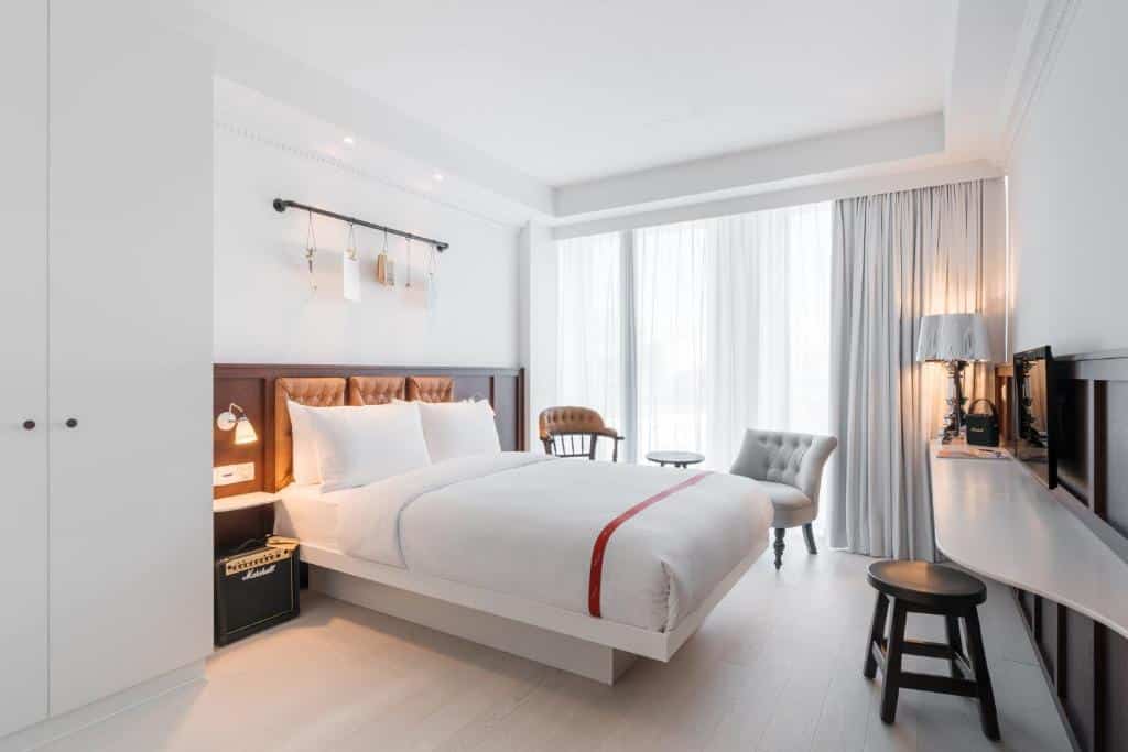 Quarto minimalista do Ruby Stella Hotel London com cama de casal, duas cadeiras, uma secretária e janelas grandes com cortinas transparentes que deixam entrar a luz.