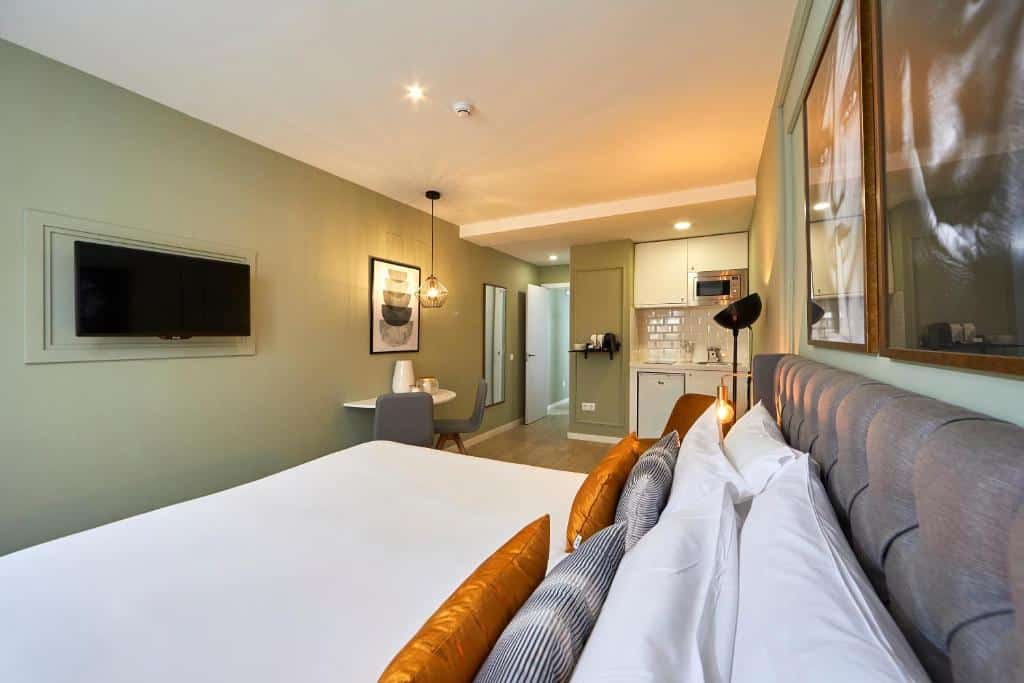 Quarto do L&H La Latina Selection, um dos melhores hotéis em Madrid. Uma cama de casal fica em frente a uma TV na parede e, ao fundo, há uma zona com micro-ondas, armários e frigorífico. Na parede da TV está uma mesa com duas cadeiras e um quadro em frente. Ao lado, um espelho está em frente à porta aberta do quarto.
