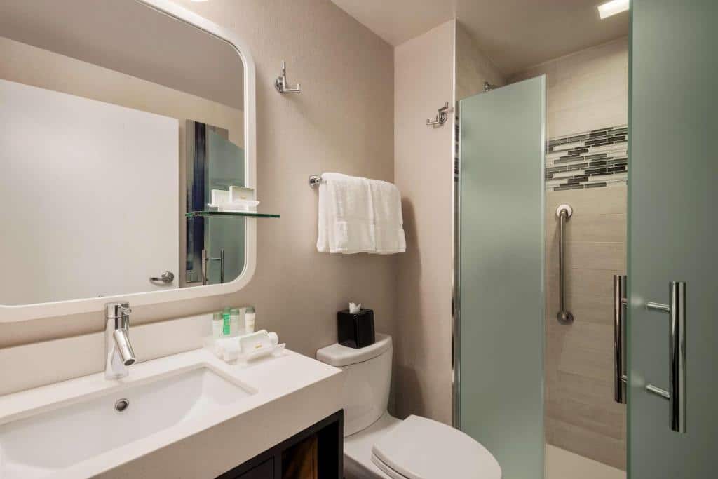 Casa de banho do Homewood Suites Midtown Manhattan Times Square South com lavatório branco e torneira moderna à esquerda e um espelho retangular com luzes embutidas. À direita, a sanita e uma cabine de duche com portas de vidro fosco; toalhas numa prateleira de metal.