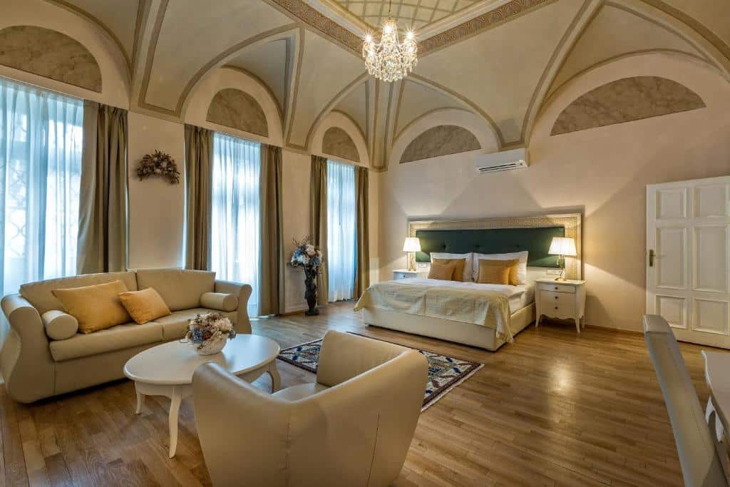 Quarto espaçoso do Little King Hotel U Malvaze, com teto abobadado, cama grande, sofás, poltronas, lustre e chão de madeira.