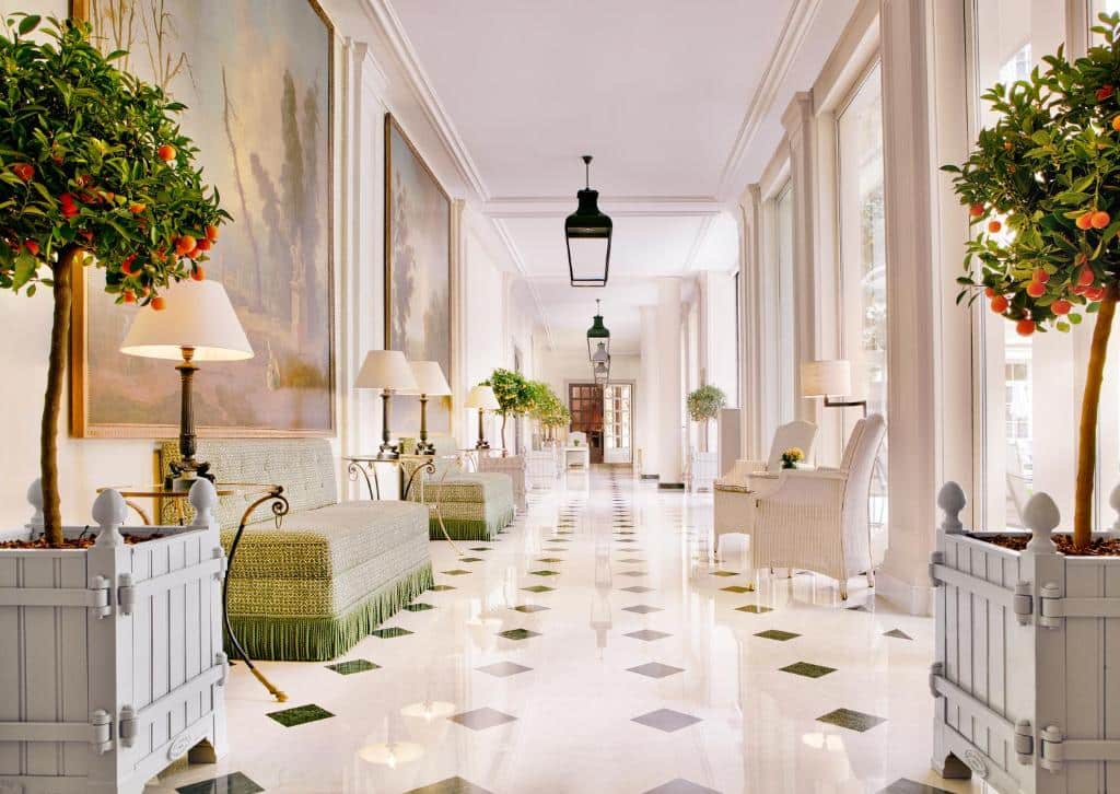 Um corredor luminoso e elegante do Le Bristol Paris - an Oetker Collection Hotel com janelas grandes, sofás verdes, vasos de laranjeiras, cadeiras e mesas de moldura branca. Este é um dos hotéis de luxo em Paris.