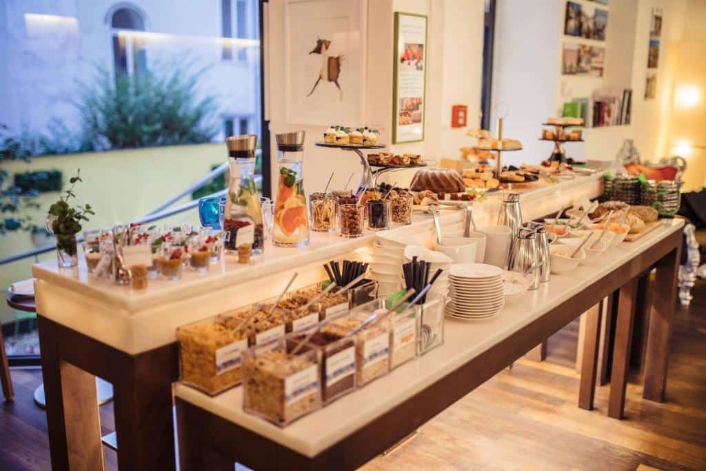 Mesa de buffet de pequeno-almoço no Hotel Rathaus, com cereais, fruta, doces, jarros de sumo, pratos e utensílios, numa sala bem iluminada.