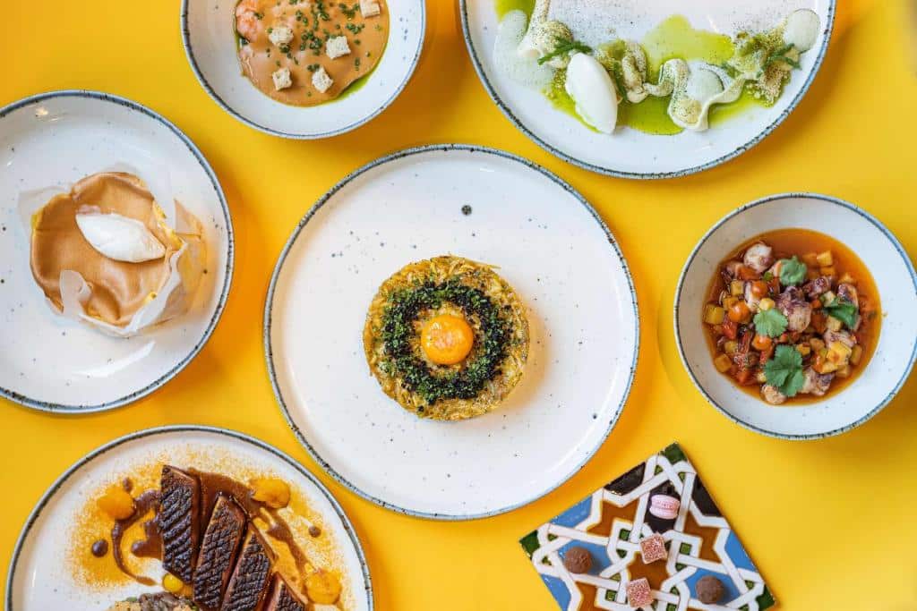 Seis pratos gourmet variados dispostos numa mesa amarela viva do art’otel amsterdam, vistos de cima.