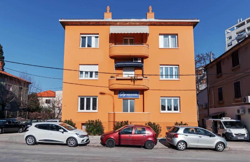 Fachada do Sky Hostel, edifício cor de laranja de três andares com carros estacionados na rua, em Kolovare