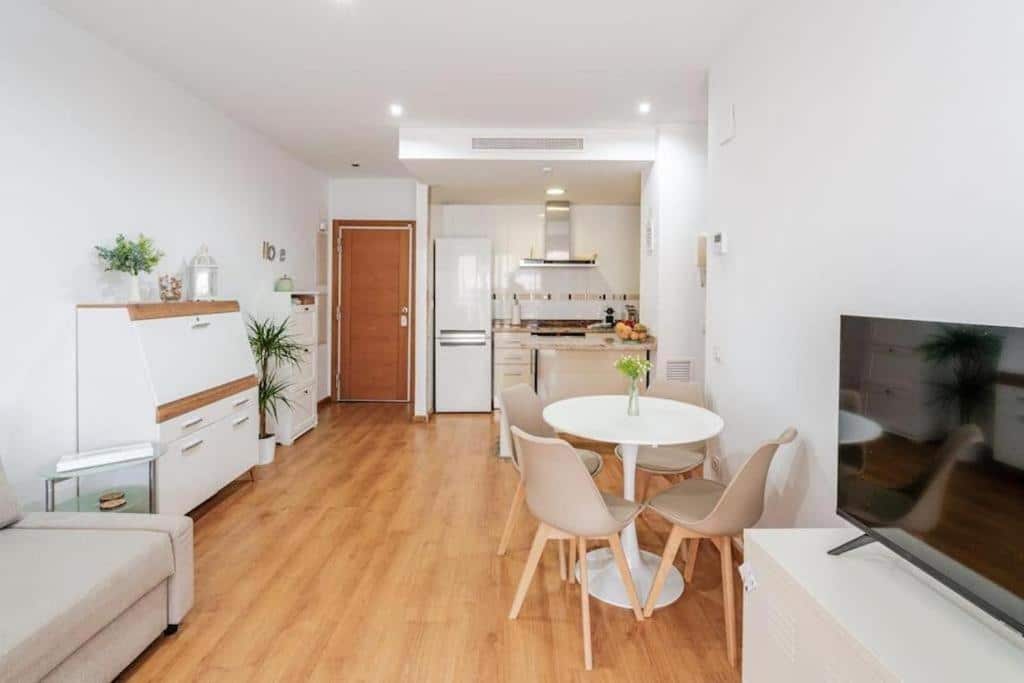 Sala de estar e cozinha do FiveStepsToSea em estilo minimalista, com móveis claros e decoração moderna, mesa redonda branca com cadeiras, frigorífico e armários brancos, pavimento de madeira e uma janela ampla com vista exterior.