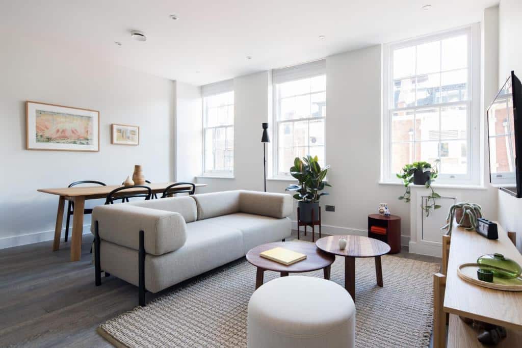 Sala de estar do apartamento Hausd – Leicester Square com um sofá de dois lugares, três janelas, uma televisão, uma mesa de centro redonda de madeira, um tapete bege, um aparador branco, uma mesa de quatro lugares e alguns vasos de plantas.