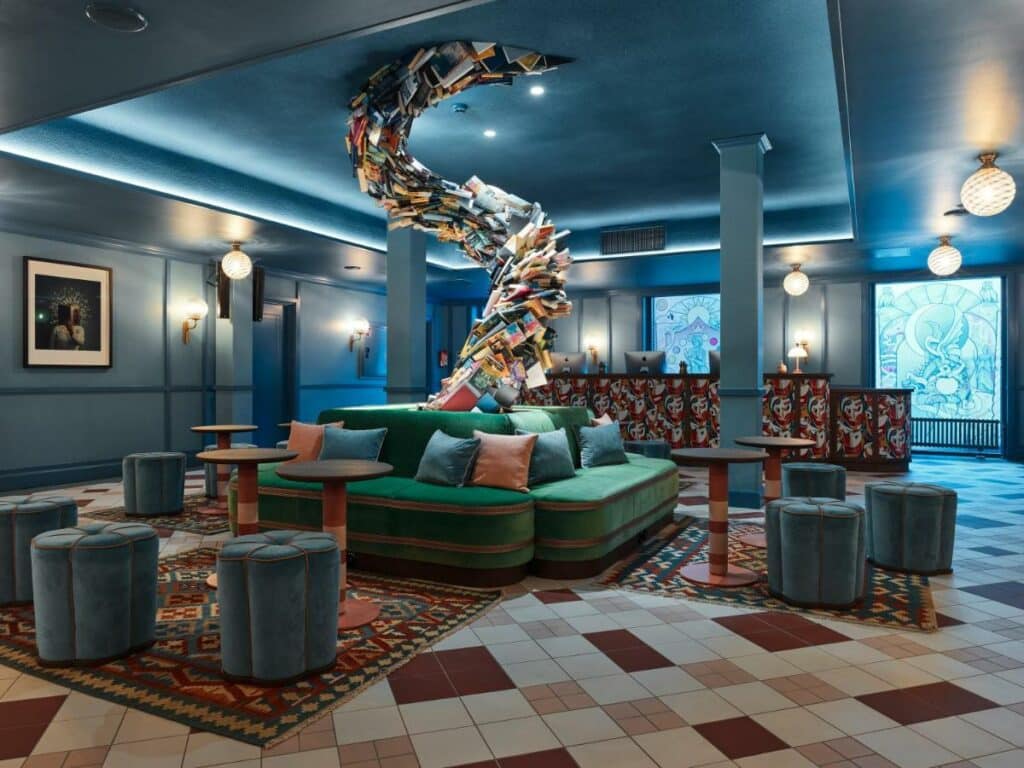 Zona comum do 25hours Hotel Indre By com vários sofás, pufes, balcão de atendimento e uma escultura feita de livros ao centro.