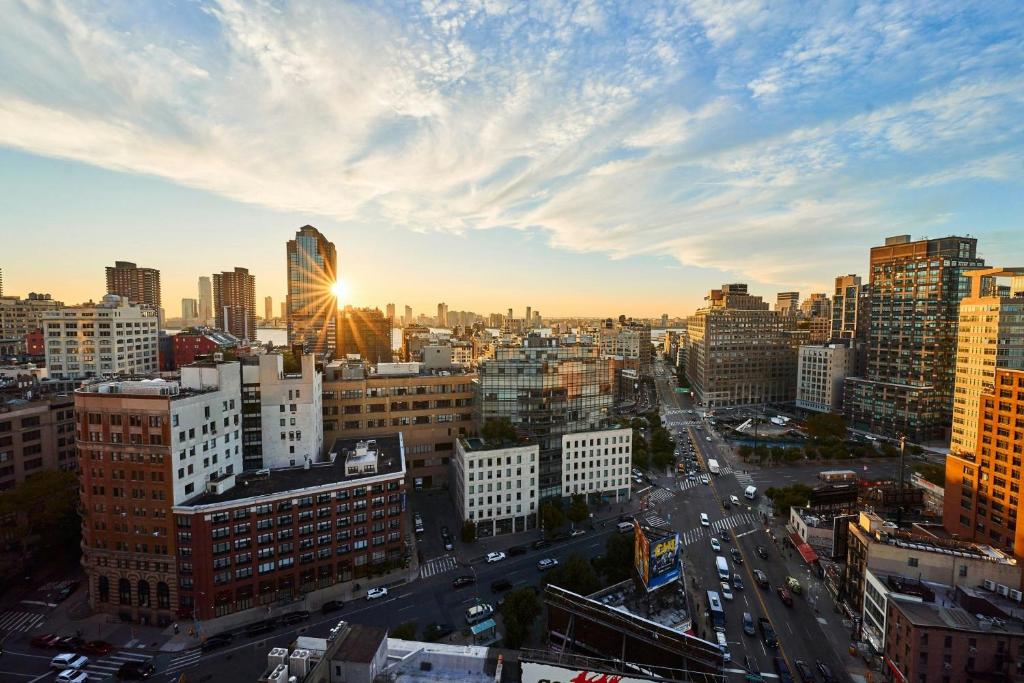 Paisagem urbana ao pôr do sol, com o sol a brilhar por entre edifícios altos e ruas movimentadas cheias de carros.