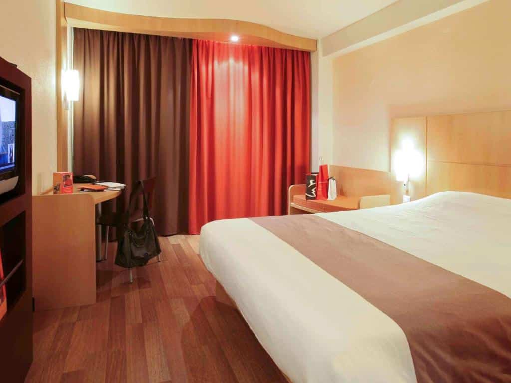 Quarto do Hotel ibis Braga com cama de casal, pavimento em madeira, pequena secretária, cadeira, TV e cortinas fechadas.