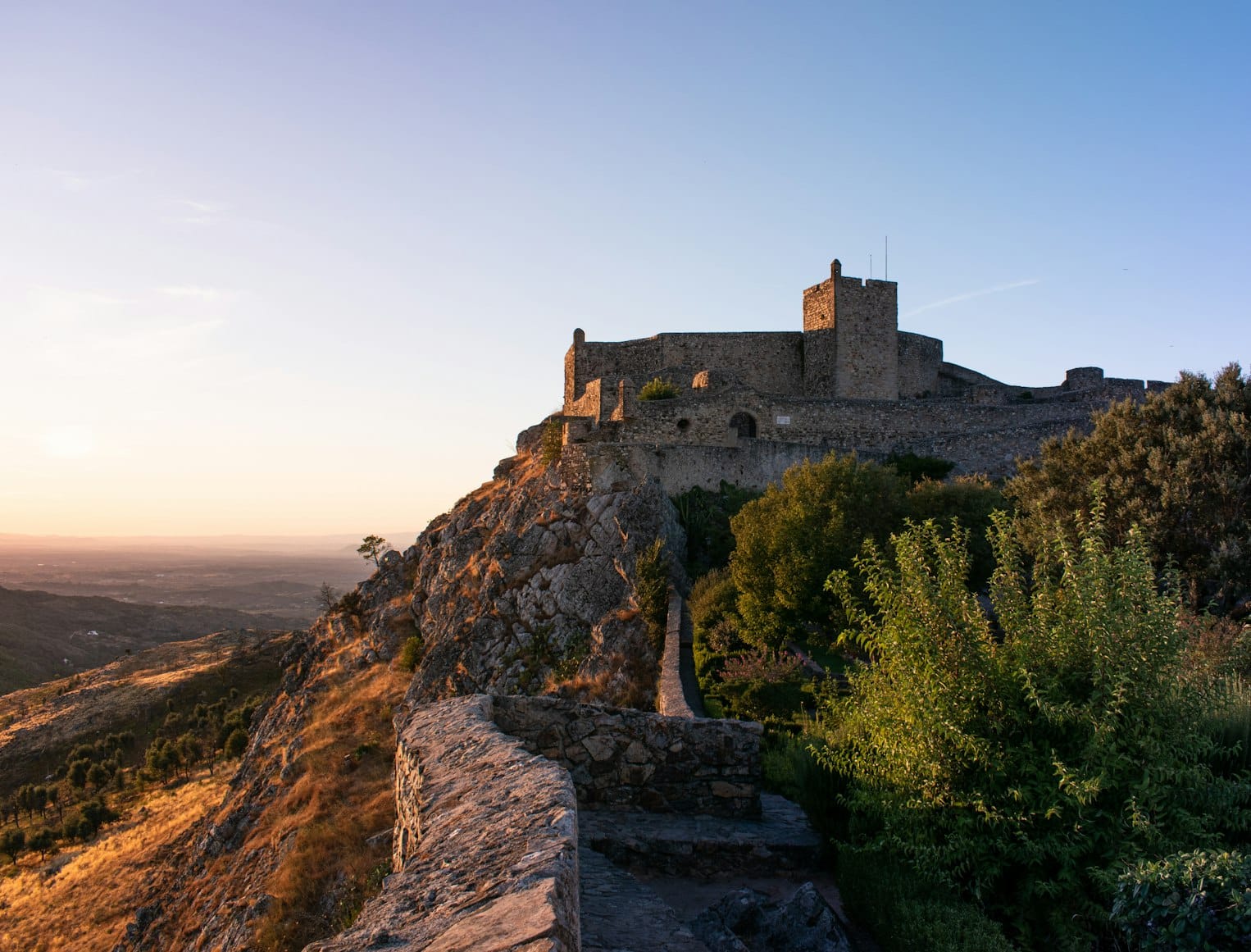 Castelo de pedra no topo de uma colina rochosa, com folhagem verde e um caminho que leva até lá, ao pôr do sol.