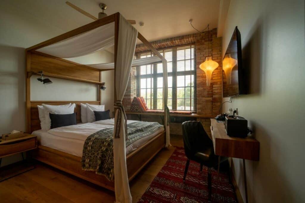 Quarto do hotel Bryggen Guldsmeden. Tem uma cama de casal com dossel, um candeeiro de luz amarela, mesa com cadeira, televisão e uma janela com um banco para descansar.