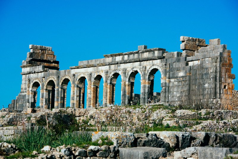 Ruínas antigas de pedra de Volubilis, Património da UNESCO, com um arco sob um céu azul claro e terreno rochoso em primeiro plano.