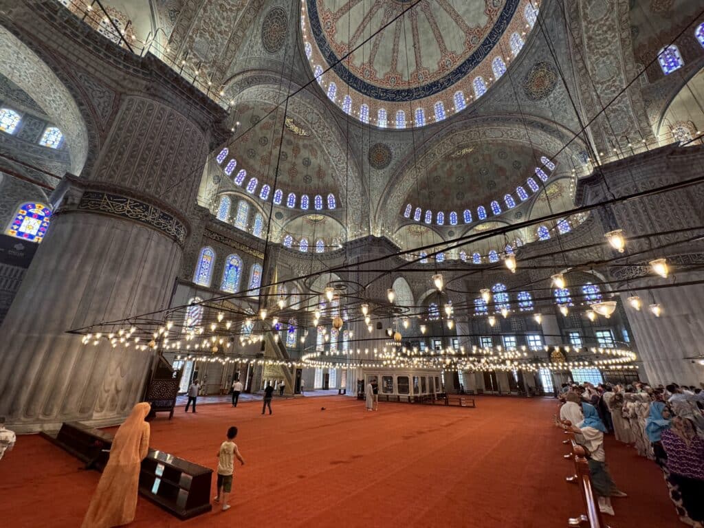 Interior de uma grande mesquita com tetos ornamentados, colunas, candeeiros suspensos e pessoas a caminhar sobre um tapete vermelho.