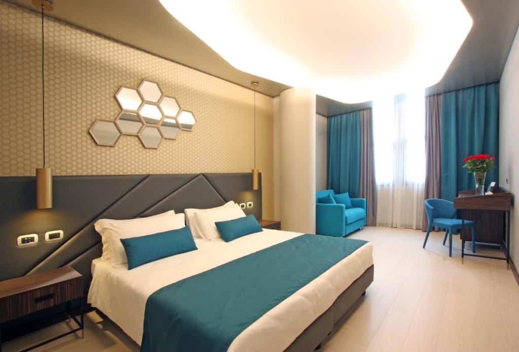 quarto do The Hive Hotel, um dos hotéis bem localizados em Roma, com cama de casal grande, mesa e candeeiro de cada lado, espelho em forma de colmeia por cima e uma zona de estar com sofá e poltrona junto a uma janela grande com cortinas, com mobiliário azul e luzes no teto