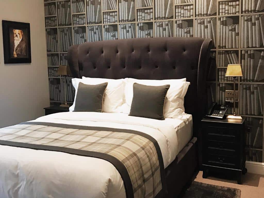 Uma cama bem feita, com almofadas cinzentas e padrão xadrez, ladeada por duas mesas de cabeceira pretas e um papel de parede com estampa de livros, no The Grafton Arms Pub & Rooms.