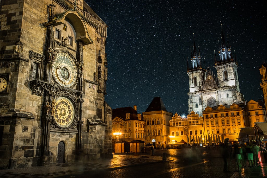 Fotografia noturna do centro histórico de Praga, com a torre do Relógio Astronómico à esquerda e, ao fundo, edifícios históricos iluminados por uma luz quente sob um céu estrelado.