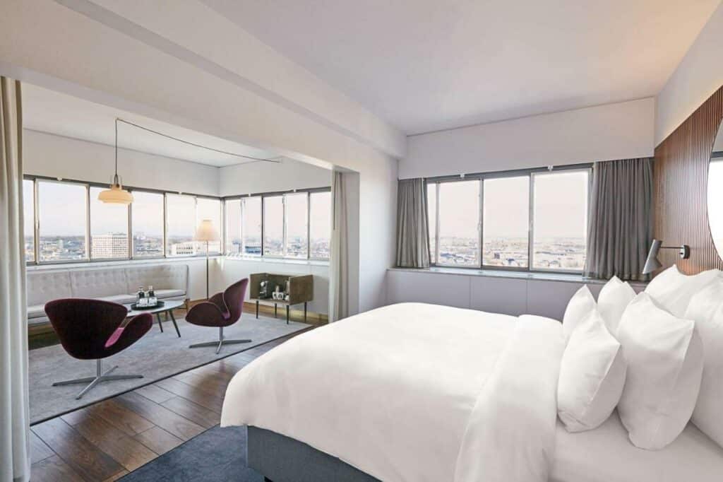 Quarto do Radisson Collection Royal Hotel com cama de casal, mesas e cadeiras e janelas panorâmicas com vista para a cidade.