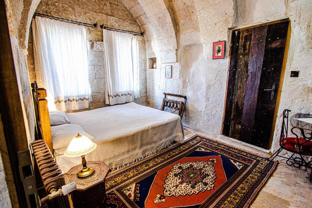 Quarto rústico do Kelebek Special Cave Hotel & Spa, um dos hotéis da Capadócia, com paredes de pedra, cama de casal, tapete colorido, porta de madeira, mesa pequena e duas janelas claras.