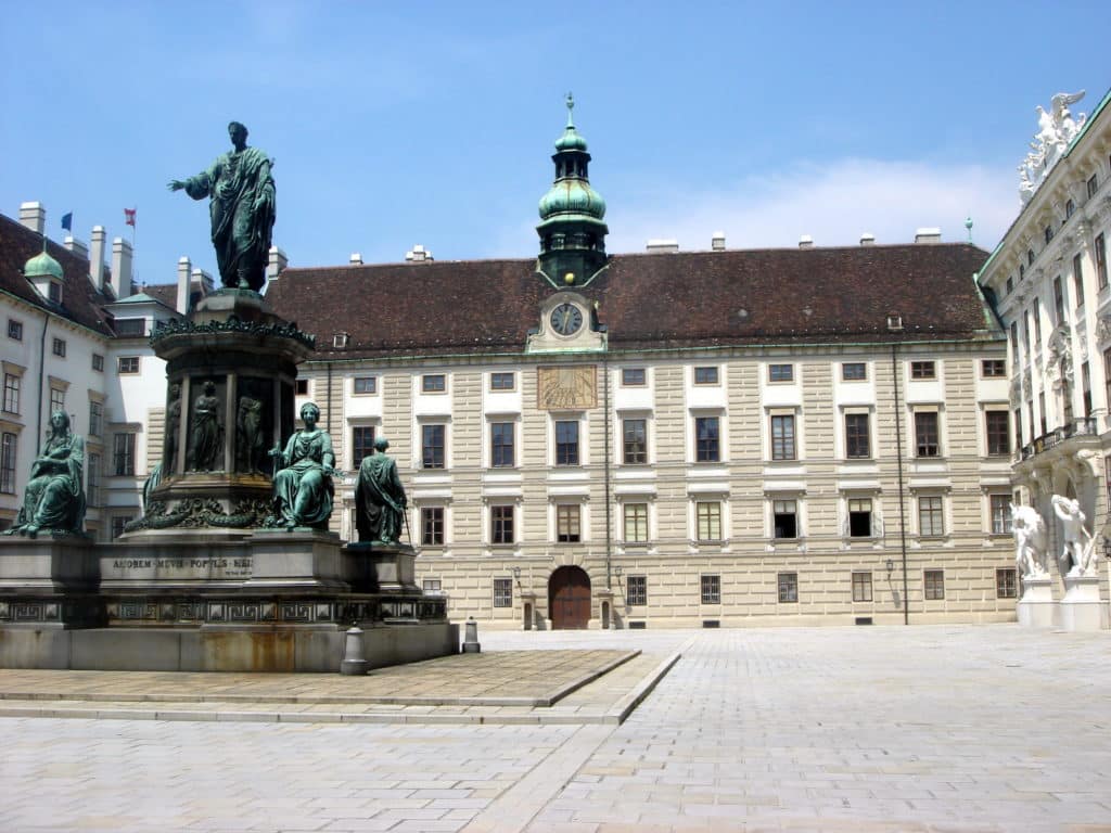 Vista exterior do Hofburg, com uma grande estátua numa praça histórica em frente a um edifício com torre de relógio, sob céu limpo.