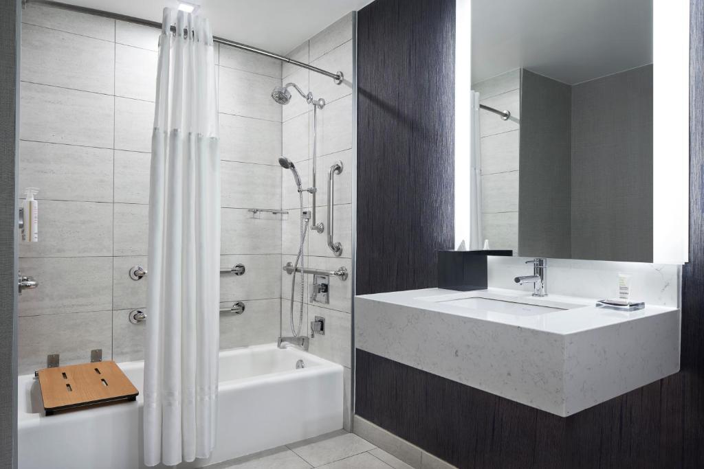 Casa de banho do New York Marriott Marquis com lavatório retangular branco e torneira moderna. À direita, um espelho grande com iluminação embutida; à esquerda, uma banheira com cortinado de duche branco.