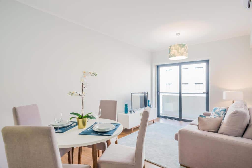 Sala de estar e jantar bem iluminada com mesa redonda, quatro cadeiras, sofá, televisão e uma grande janela para uma varanda, no 137 LuxResidence.