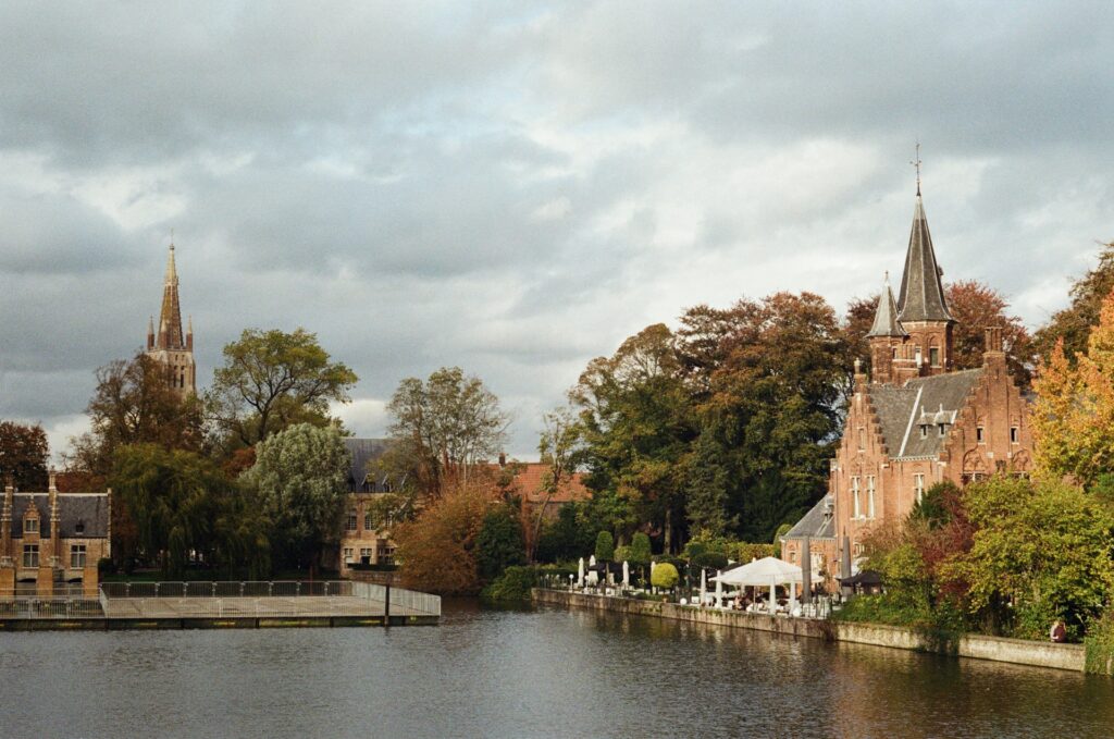 Paisagem de Bruges, na Bélgica, mostrando o canal rodeado por edifícios históricos de tijolo vermelho e torres pontiagudas. À esquerda, uma igreja gótica com uma torre alta e fina destaca-se no cenário, enquanto à direita há uma construção com torres menores, também medieval. As árvores de outono acrescentam toques de laranja e verde ao cenário, e um pequeno cais com mesas e cadeiras oferece um local relaxante à beira da água.
