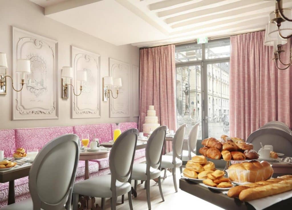Sala de pequeno-almoço bem iluminada do La Maison Favart, com decoração cor-de-rosa, mesas postas e um buffet junto a grandes janelas.