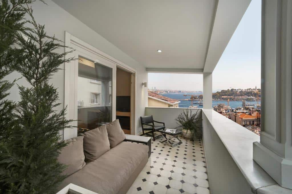 Uma varanda coberta do The Story Hotel Pera com um sofá, cadeiras, vasos de plantas e uma vista da cidade e da água ao longe.