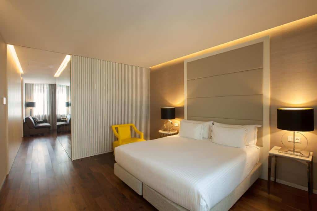 Quarto moderno no BessaHotel Liberdade com uma cama grande, cadeira amarela, dois candeeiros pretos, pavimento em madeira e uma zona de estar ao fundo.