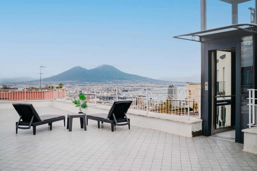 Terraço com vista panorâmica sobre Nápoles e o imponente Monte Vesúvio ao fundo, numa tarde solarenga, com duas espreguiçadeiras pretas de design moderno sobre o pavimento de ladrilhos claros, no Vista Napoli Residence by Casa Napoletana.