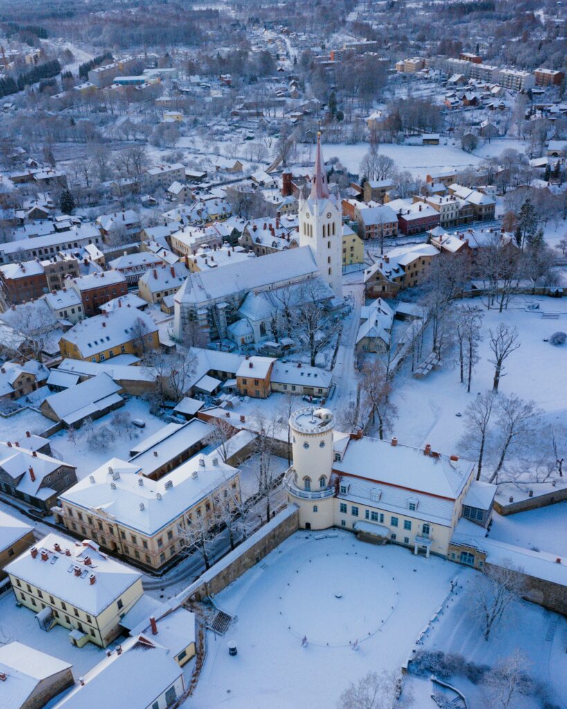 Vista aérea de Cēsis, na Letónia, coberta de neve durante o inverno, com casas e ruas rodeadas por árvores despidas. No centro, destaca-se uma grande igreja com torre alta e pontiaguda.