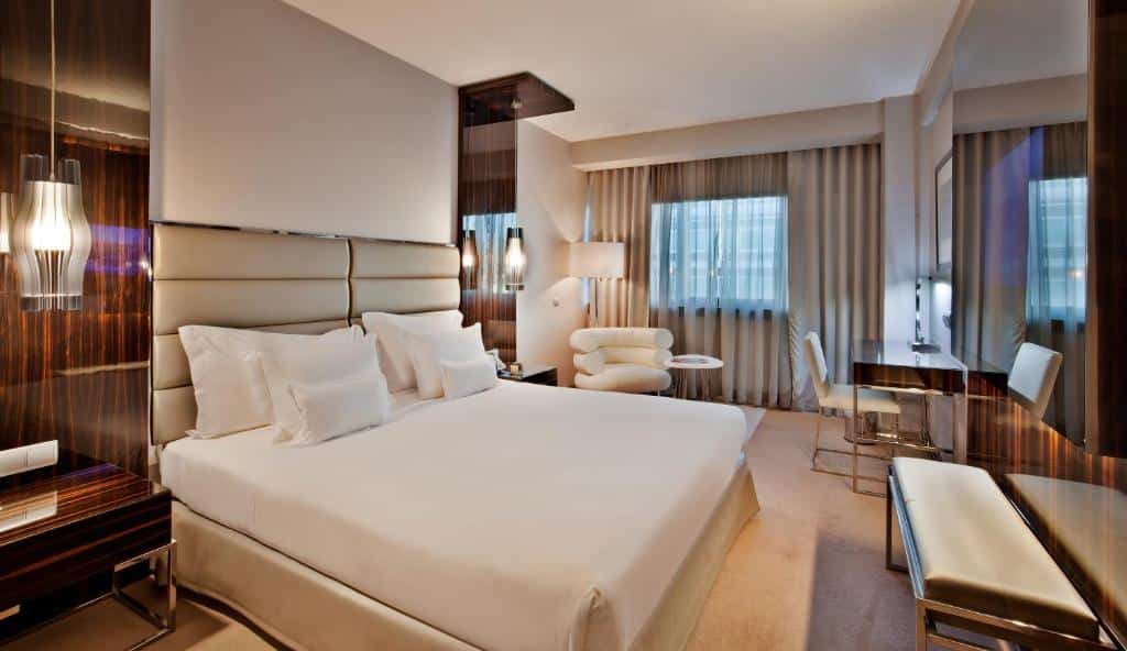 Quarto moderno no Altis Grand Hotel com cama grande, lençóis brancos, zona de estar, secretária e janelas amplas com cortinas.