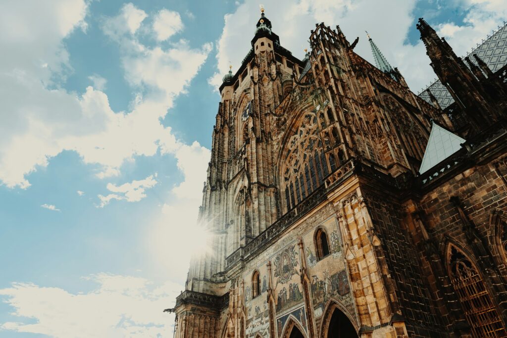 Uma grande catedral gótica com detalhes ornamentados destaca-se num céu azul brilhante, com o sol a brilhar por trás do edifício.
