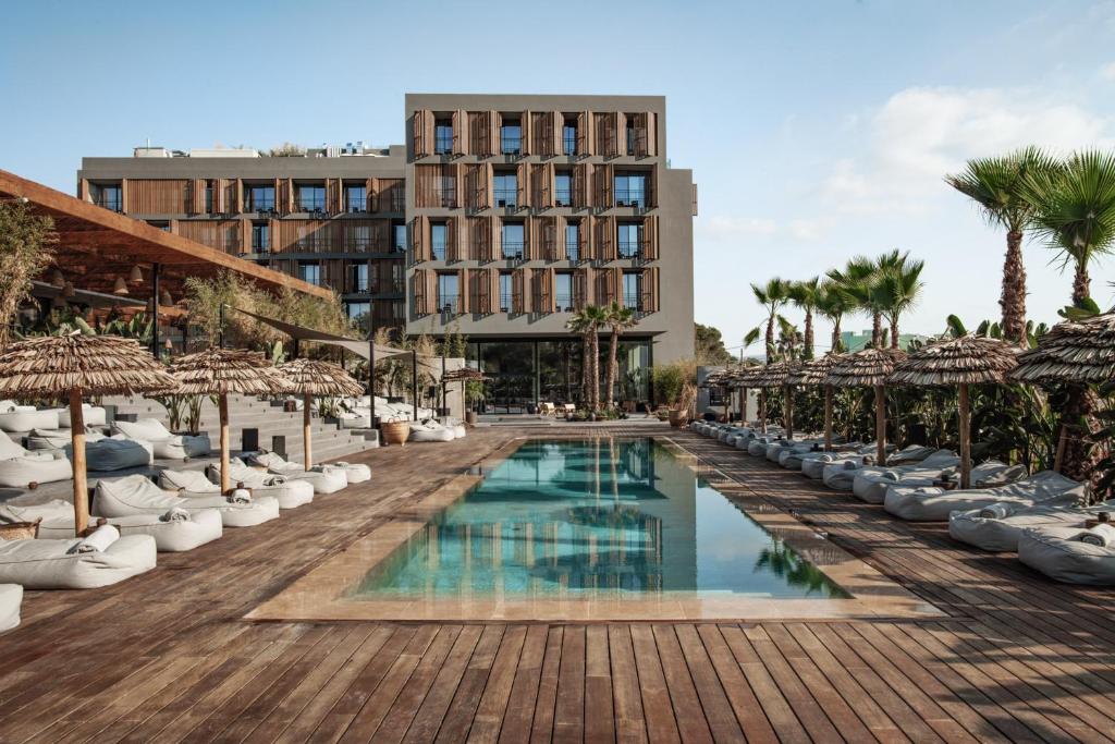 Zona exterior do OKU Ibiza em estilo moderno, com detalhes em madeira e uma piscina retangular rodeada de espreguiçadeiras e chapéus de sol de palha.