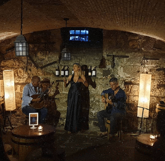A Casa do Fado, com três músicos a atuar num ambiente rústico e de luz suave, com paredes de pedra e garrafas de vinho.