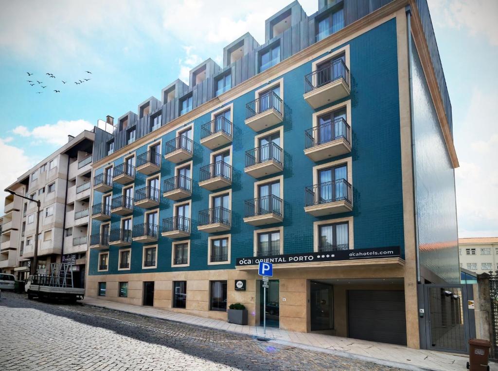 Edifício moderno do Oca Oriental Porto Hotel de cinco andares com pequenas varandas e fachada azul, numa rua de calçada.
