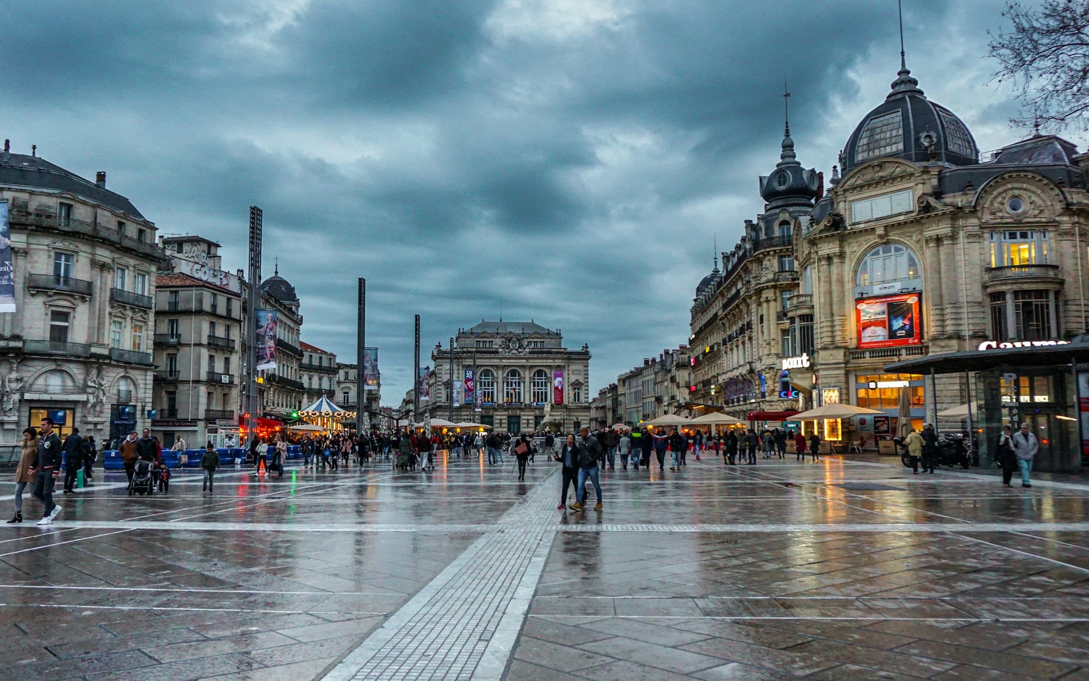 Praça movimentada com pessoas a caminhar, rodeada de edifícios ornamentados, com o pavimento molhado da chuva, em Montpellier.