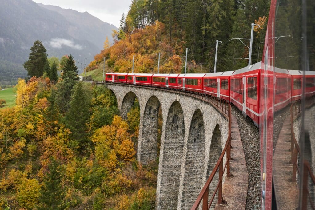 Um comboio vermelho em Albula percorre um viaduto de pedra curvo, rodeado de folhagem de outono e com montanhas ao fundo. Representa como viajar de comboio pela Suíça.