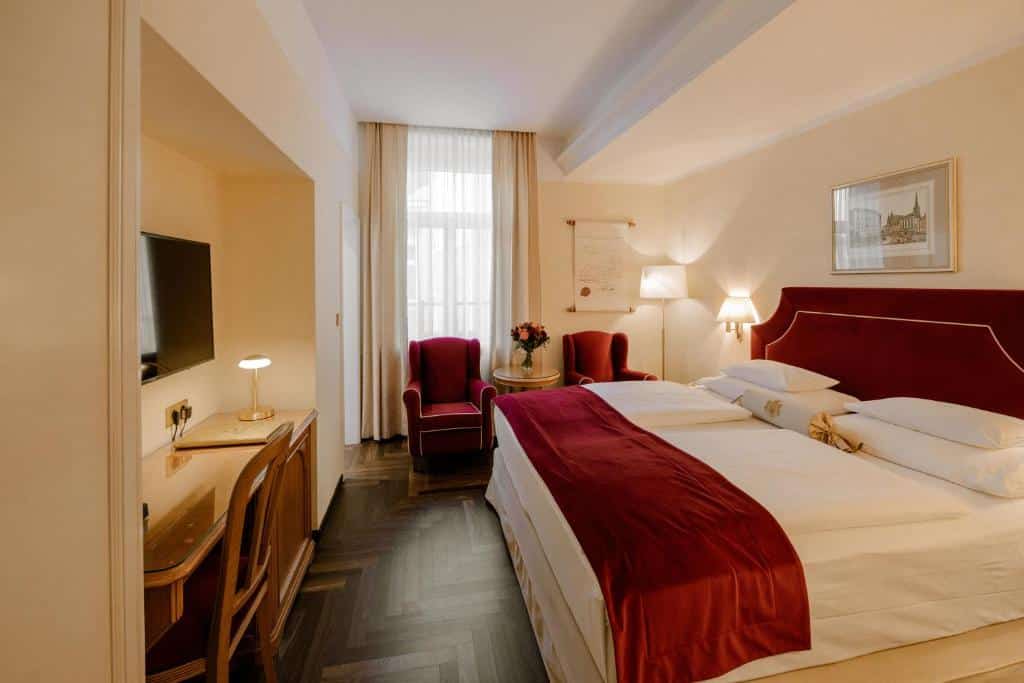 Quarto no Hotel Erzherzog Rainer, com cama de casal, apontamentos em vermelho, duas poltronas, uma TV sobre uma mesa e uma janela com cortinados transparentes.
