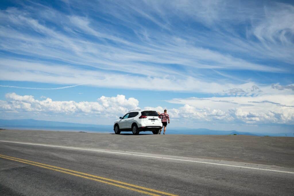 Carro branco parado numa estrada, com uma pessoa de pé do lado direito, em dia de céu claro repleto de nuvens.