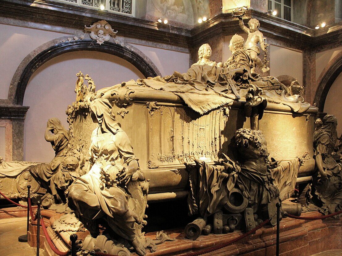 Sarcófago de pedra na Cripta Imperial, ornamentado com figuras esculpidas, numa sala pouco iluminada.