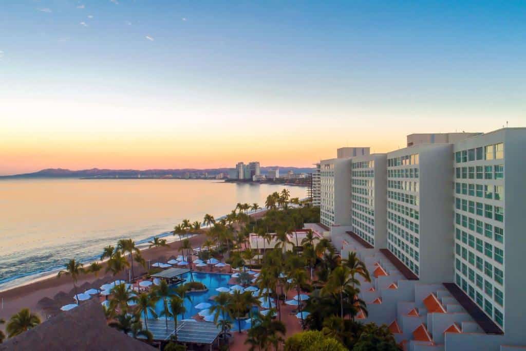 Hotel alto à beira-mar com piscinas e palmeiras ao pôr do sol, com vista para o oceano calmo e a paisagem urbana distante.