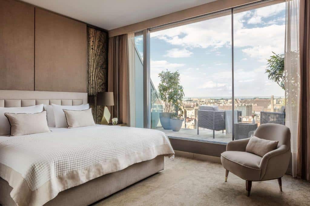 Um quarto do Anantara New York Palace com uma cama grande, poltrona e janela do chão ao teto, com vista para um terraço e para o horizonte da cidade.