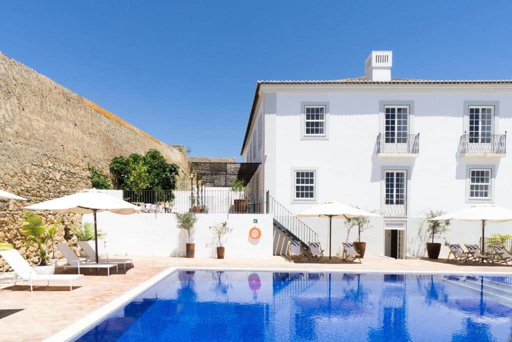 Uma piscina exterior retangular com água azul em frente a um edifício branco de dois andares, com varandas e chapéus de sol à beira da piscina, na Casa Mãe em Lagos.