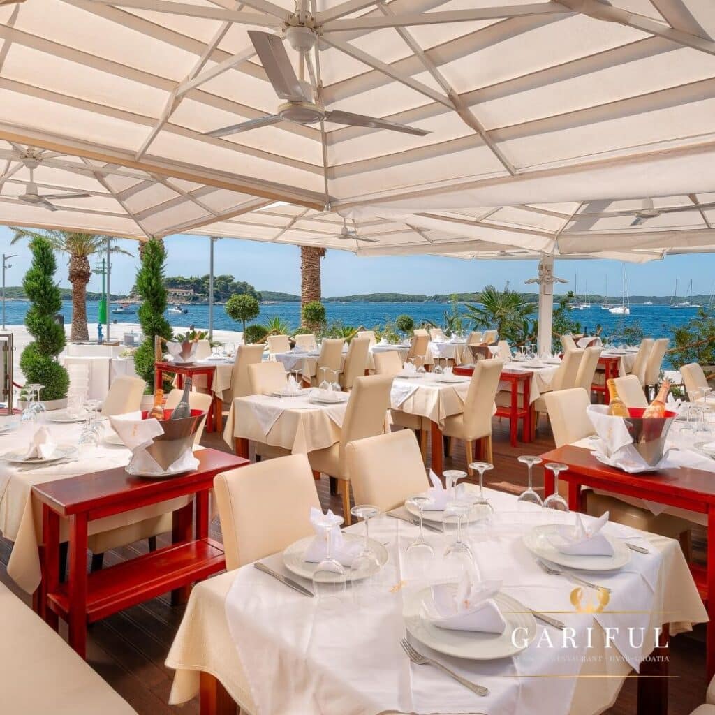 Restaurante Gariful, em Hvar, ao ar livre, com mesas sob toldos brancos e vista para uma marginal pitoresca com palmeiras e barcos.