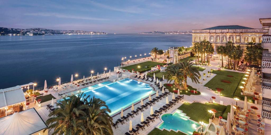 Çırağan Palace Kempinski Istanbul à beira-mar, com piscina exterior iluminada, jardins, palmeiras, espreguiçadeiras e vista para o mar ao anoitecer.