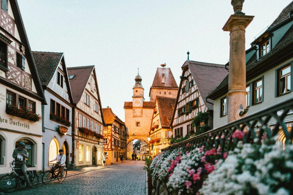 Cenário de uma rua em Rothenburg ob der Tauber, Alemanha, conhecida pela arquitetura em estilo enxaimel. A rua é ladeada por construções de fachadas decoradas e janelas com floreiras coloridas. A rua é ainda adornada com uma balaustrada cheia de flores. Ao fundo, a torre do relógio ergue-se como parte da porta da cidade.